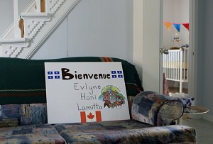 Photo : sur un divan vert, dans une maison aux murs blancs, une affiche blanche Bienvenue Evlyne, Hani et Lamitta. Drapeaux du Québec haut et un du Canada au bas.