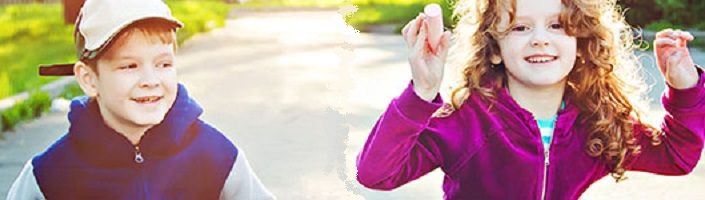 Deux enfants souriants, un garçon et une fille, sur une rue. Les deux ont les cheveux roux. Il porte une casquette de baseball. Elle porte les cheveux long frisés.
