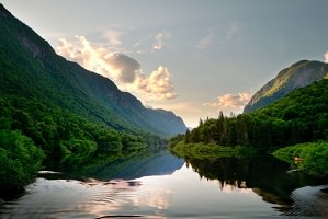 Photo officielle : rivière calme dans une vallée très verte, forêt de conifères, montagnes non loin et quelques nuages. Soleil derrière les montagnes.