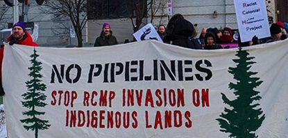 Photo d'une manif sur une voie ferrée, l'hiver, avec bannière beige « No pipelines - Stop RCMP invasion on indigenous lands ». Dessin de deux sapins verts sur la bannière. Foule derrière.