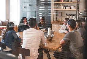 Photo de sept jeunes adultes autour d'une grande table en bois dans un café ensoleillé.