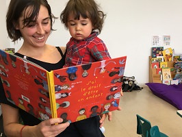 Photo d'une jeune femme tenant un-e gamin-e d'environ 2-3 ans, aux cheveux longs bruns ébouriffés un peu et chemise rouge; et tenant aussi un grand livre rouge avec plusieurs personnages dessinés dessus.